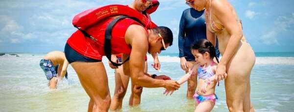 A princípio, a identificação da criança na praia é extremamente importante para garantir sua segurança e evitar que se percam. Nesse sentido, quando as crianças estão na praia, elas podem se distrair facilmente e se afastar dos pais ou responsáveis, o que pode ser perigoso e assustador para elas. Ainda mais que, ao identificar a criança, os pais ou responsáveis podem ser rapidamente localizados em caso de emergência ou se a criança se perder. Como também, existem várias formas de identificar a criança na praia, como colocar uma pulseira com informações de contato dos pais ou responsáveis, escrever as informações com caneta impermeável no braço da criança, ou mesmo vestir a criança com uma camiseta personalizada. Além disso, é importante orientar a criança sobre como agir caso se perca na praia, como procurar um salva-vidas ou uma autoridade local e pedir ajuda. Também é importante que os pais ou responsáveis estejam sempre atentos e observando a criança de perto enquanto estiverem na praia. Coloque uma pulseira de identificação Ou seja, esta medida é fundamental para caso a criança se perca. "identificar sua criança com uma pulseirinha faz toda a diferença. Em segundos, uma criança que estava próxima some e você não acha mais. Escreva o nome da criança, dos pais e um telefone para contato", ensina o major Fernando Chailon Rodrigues Fonteles da 1ª Companhia de Salvamento Marítimo do Batalhão de Busca e Salvamento. "Também é fundamental dar um ponto de referência para a criança, como, por exemplo, uma placa ou um quiosque, avisando que, caso ela se perca, fique lá até alguém ir buscá-la", completa o guarda-vidas do Corpo de Bombeiros do Ceará. Supervisione a criança de perto Como também, os perigos da praia não são poucos - a criança pode se perder, entrar sozinha no mar, colocar objetos indevidos na boca etc. "Crianças não tem medo de nada, acham que nada é perigoso. Essa é a idade que elas ainda estão descobrindo tudo", observa o guarda-vidas. Por isso, os pais/responsáveis devem supervisionar todas as brincadeiras de perto.